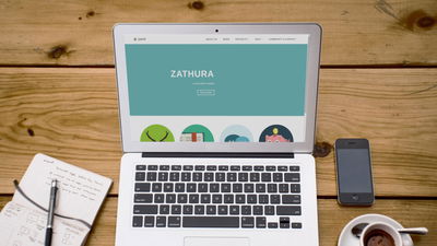 A photograph of a laptop on a table along a cup of coffee, smartphone and notepad.